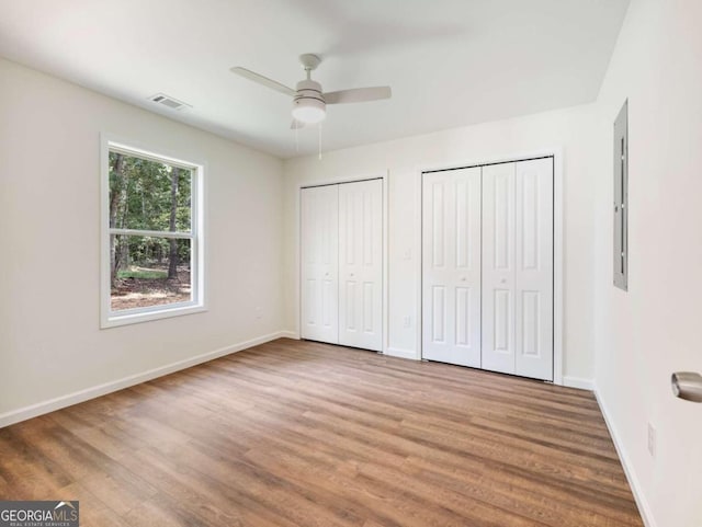 unfurnished bedroom with electric panel, ceiling fan, wood-type flooring, and two closets