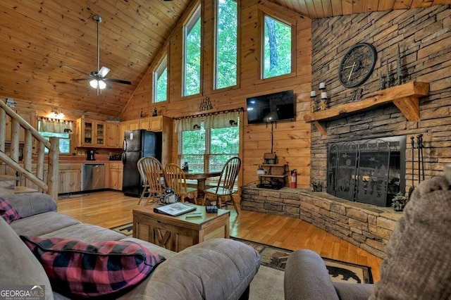 living room featuring ceiling fan, wooden walls, light wood-type flooring, high vaulted ceiling, and a stone fireplace