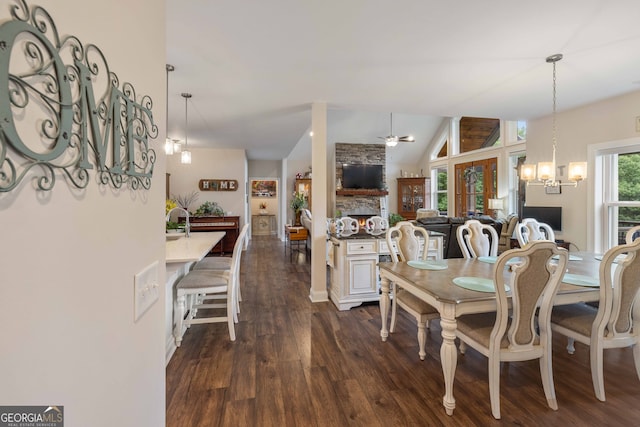 dining area featuring ceiling fan with notable chandelier, a stone fireplace, vaulted ceiling, and dark wood-type flooring