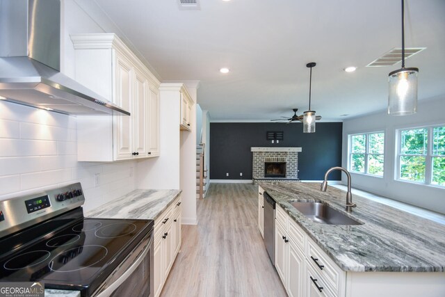 kitchen with tasteful backsplash, wall chimney range hood, appliances with stainless steel finishes, stone countertops, and a brick fireplace