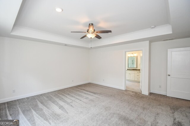 unfurnished bedroom featuring a raised ceiling, light colored carpet, ensuite bath, and ceiling fan