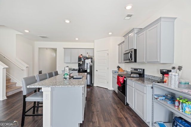 kitchen with dark wood-type flooring, stainless steel appliances, a kitchen breakfast bar, light stone countertops, and a center island with sink