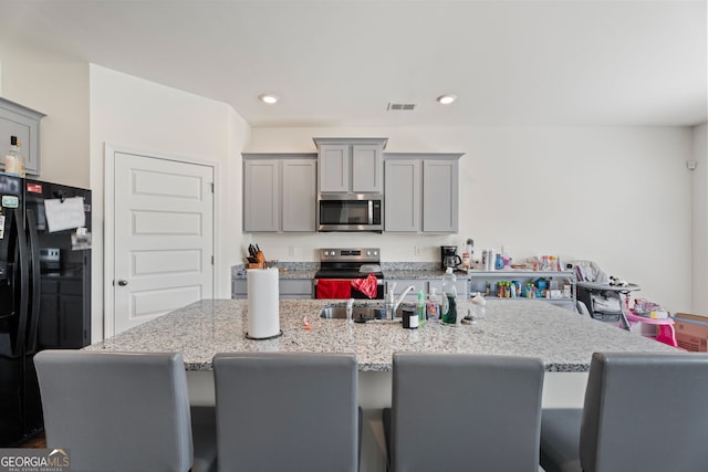 kitchen featuring light stone counters, stainless steel appliances, a center island with sink, and gray cabinetry