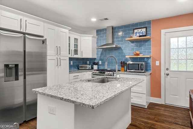 kitchen with wall chimney range hood, dark hardwood / wood-style flooring, decorative backsplash, appliances with stainless steel finishes, and a kitchen island with sink