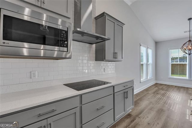 kitchen with stainless steel microwave, black electric cooktop, light wood-type flooring, wall chimney exhaust hood, and vaulted ceiling