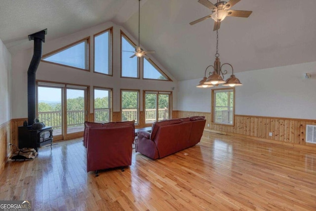 living room featuring light wood-type flooring, high vaulted ceiling, ceiling fan with notable chandelier, and a wood stove