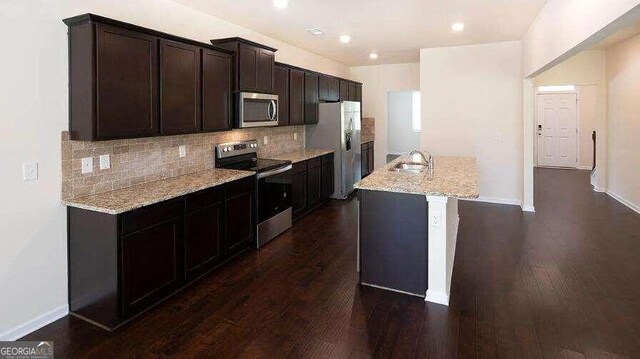 kitchen featuring dark hardwood / wood-style flooring, stainless steel appliances, an island with sink, backsplash, and light stone counters