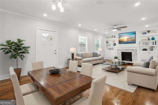 dining area featuring a stone fireplace, wood-type flooring, built in features, ceiling fan, and ornamental molding