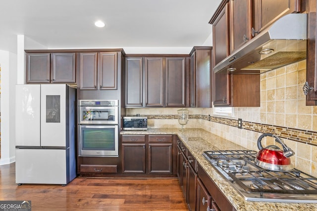 kitchen with wall chimney range hood, backsplash, dark wood-type flooring, appliances with stainless steel finishes, and light stone counters