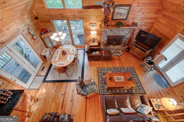 living room with a stone fireplace, high vaulted ceiling, wooden walls, ceiling fan, and hardwood / wood-style flooring