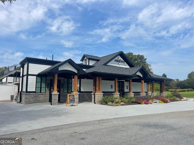 view of front of house featuring driveway, stone siding, covered porch, and roof with shingles