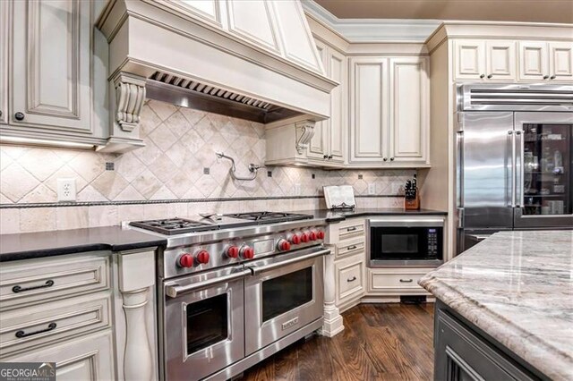 kitchen with dark stone counters, built in appliances, premium range hood, dark wood-type flooring, and tasteful backsplash