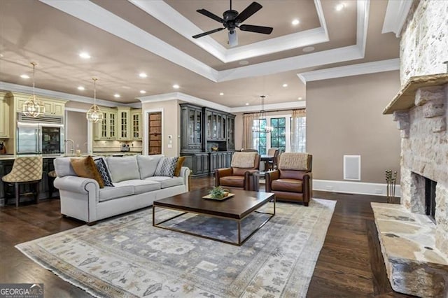 living room featuring a stone fireplace, dark hardwood / wood-style flooring, ceiling fan with notable chandelier, a raised ceiling, and crown molding