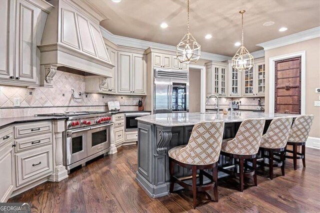 kitchen with dark stone counters, custom range hood, dark hardwood / wood-style floors, a center island with sink, and premium appliances