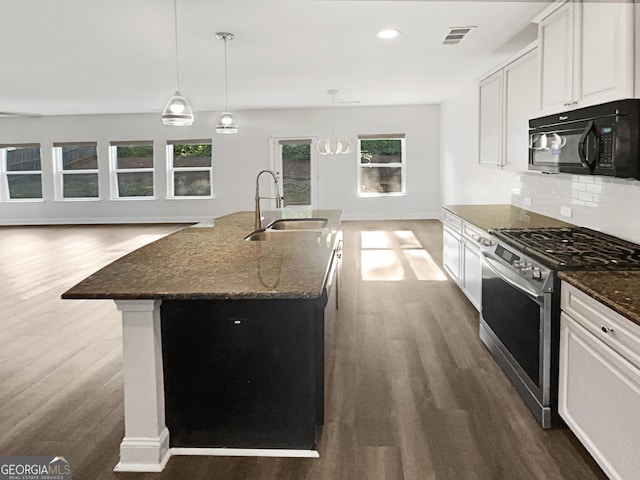 kitchen with black microwave, white cabinetry, stainless steel range with gas cooktop, dark stone counters, and an island with sink