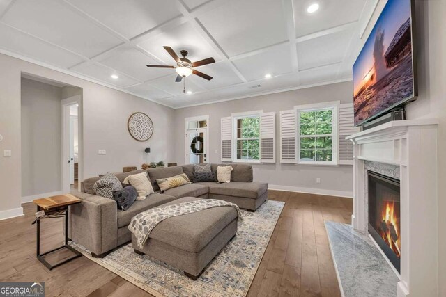 living room featuring ceiling fan, hardwood / wood-style flooring, a premium fireplace, coffered ceiling, and ornamental molding