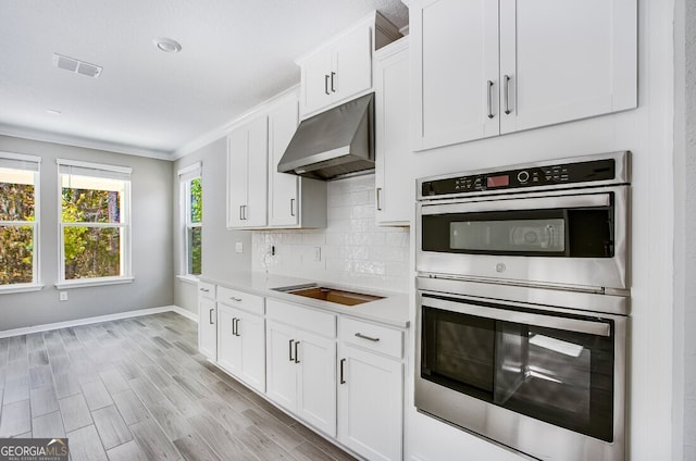 kitchen with stainless steel double oven, stovetop, white cabinets, light hardwood / wood-style floors, and backsplash