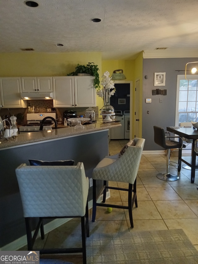 kitchen with white gas stove, light tile patterned flooring, under cabinet range hood, separate washer and dryer, and white cabinetry