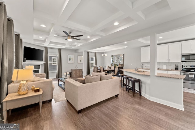 living room featuring coffered ceiling, ceiling fan with notable chandelier, hardwood / wood-style floors, sink, and beamed ceiling