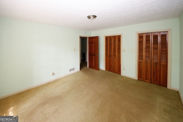 unfurnished bedroom with a textured ceiling, light carpet, visible vents, baseboards, and two closets