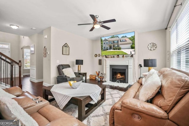 living room featuring a warm lit fireplace, baseboards, ceiling fan, dark wood-type flooring, and stairs