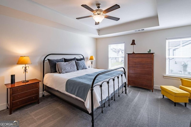 bedroom with dark colored carpet, a raised ceiling, ceiling fan, and multiple windows