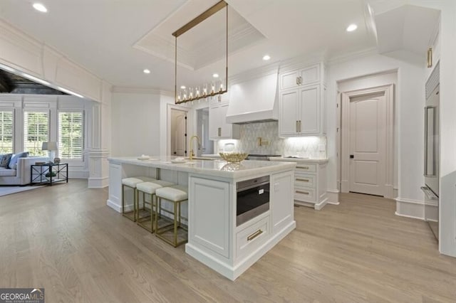 kitchen featuring light hardwood / wood-style flooring, custom exhaust hood, a center island with sink, and a raised ceiling