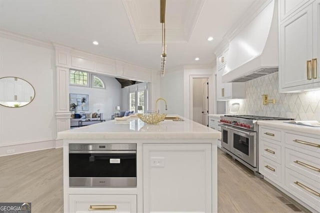 kitchen featuring custom range hood, decorative backsplash, an island with sink, light hardwood / wood-style floors, and stainless steel appliances