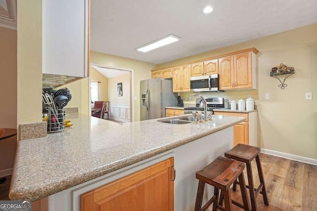 kitchen with kitchen peninsula, appliances with stainless steel finishes, a breakfast bar, light brown cabinetry, and light hardwood / wood-style floors