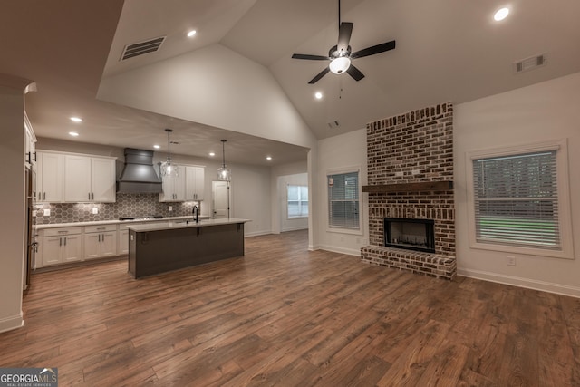 kitchen featuring a brick fireplace, dark hardwood / wood-style flooring, premium range hood, decorative light fixtures, and a center island with sink