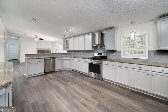 kitchen with ceiling fan, wall chimney exhaust hood, wood-type flooring, appliances with stainless steel finishes, and a textured ceiling