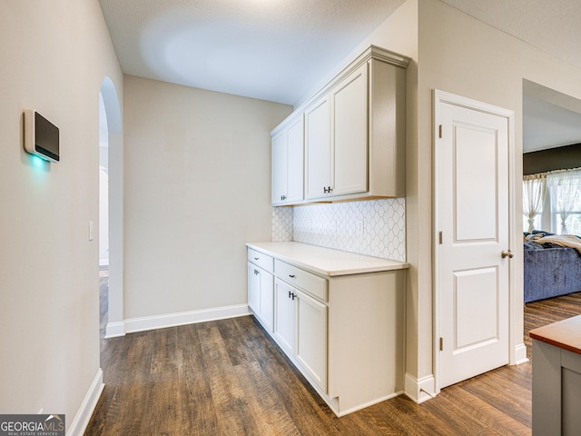 kitchen with arched walkways, dark wood-type flooring, light countertops, white cabinetry, and backsplash