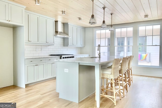 kitchen featuring a center island with sink, hanging light fixtures, wall chimney range hood, and light hardwood / wood-style floors