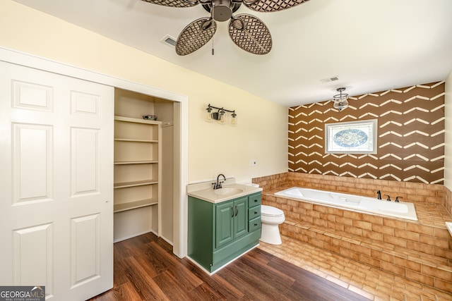 bathroom with hardwood / wood-style floors, tiled bath, vanity, ceiling fan, and toilet