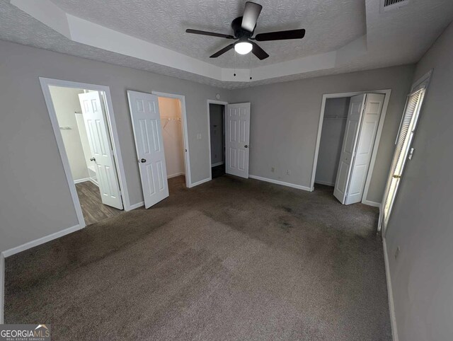 unfurnished bedroom featuring a raised ceiling, ceiling fan, a textured ceiling, ensuite bath, and dark colored carpet