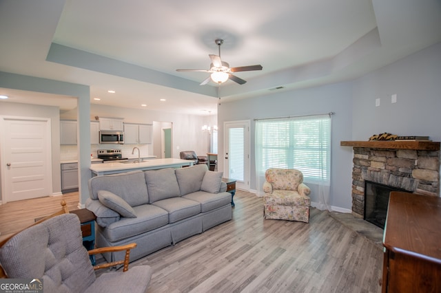 living room featuring light wood-type flooring, a raised ceiling, a stone fireplace, and ceiling fan