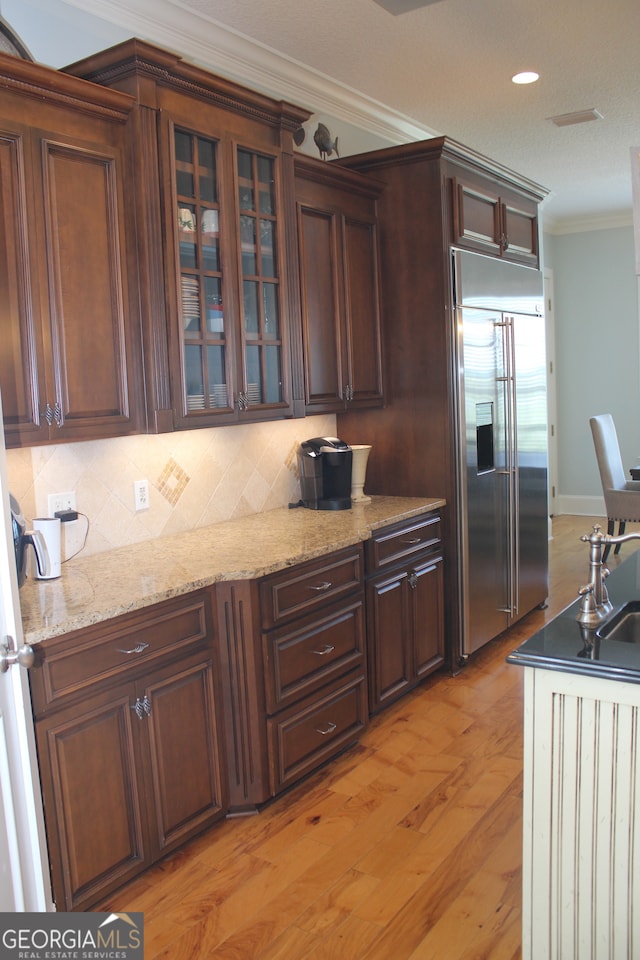 kitchen with stainless steel built in fridge, light stone counters, ornamental molding, backsplash, and light wood-type flooring