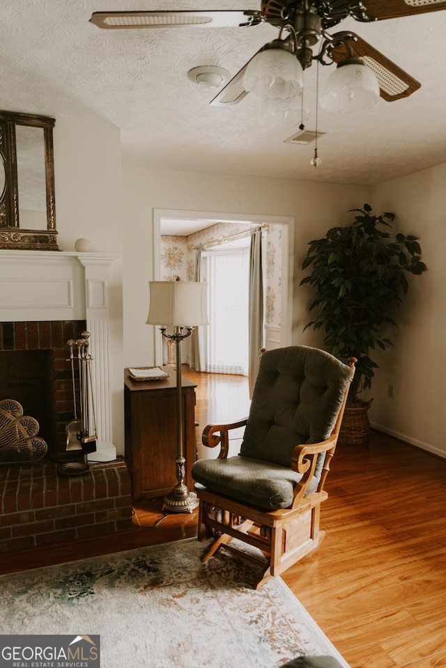 sitting room with a brick fireplace, a textured ceiling, wood-type flooring, and ceiling fan