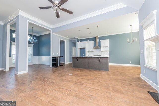 kitchen with white cabinetry, tasteful backsplash, an island with sink, wall chimney range hood, and ceiling fan with notable chandelier