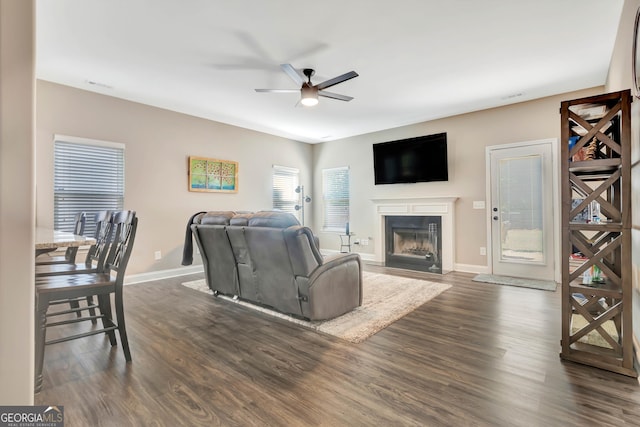 living area with dark wood-style floors, a fireplace with flush hearth, baseboards, and a ceiling fan