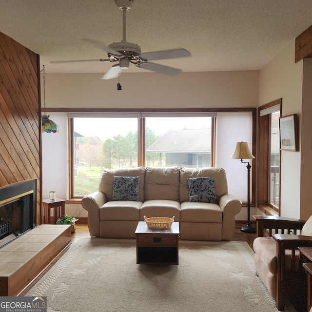 living room with a tiled fireplace, ceiling fan, a textured ceiling, and wood walls