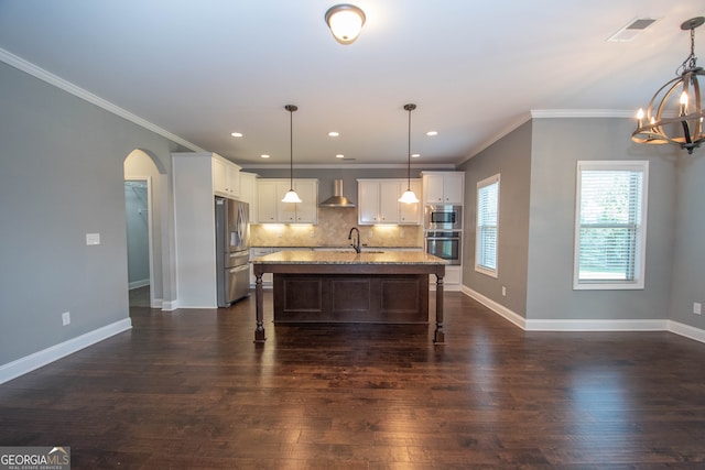 kitchen featuring a center island with sink, wall chimney range hood, appliances with stainless steel finishes, decorative light fixtures, and dark hardwood / wood-style flooring