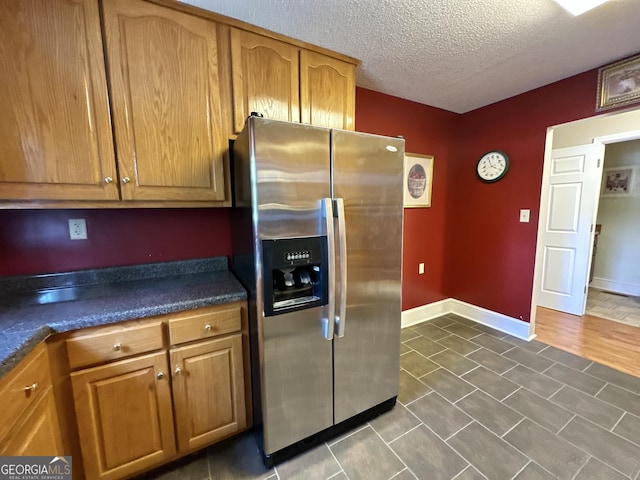 kitchen with a textured ceiling, baseboards, stainless steel refrigerator with ice dispenser, brown cabinetry, and dark countertops