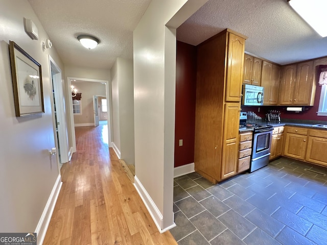 kitchen featuring a textured ceiling, baseboards, appliances with stainless steel finishes, brown cabinetry, and dark countertops