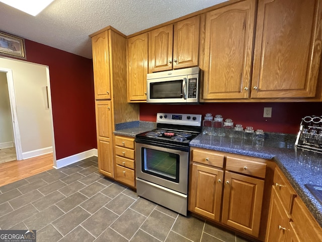 kitchen with baseboards, dark stone counters, appliances with stainless steel finishes, brown cabinets, and a textured ceiling