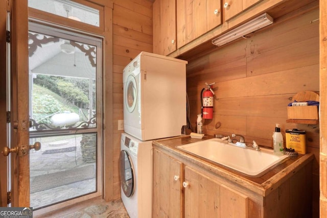 clothes washing area with wood walls, stacked washer and clothes dryer, sink, and cabinets