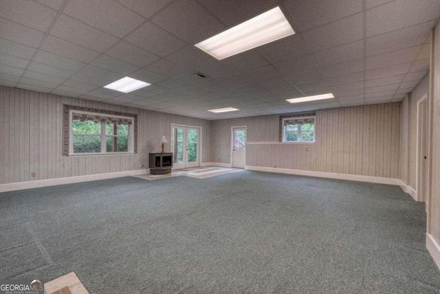 basement featuring a drop ceiling, carpet, wood walls, and a wood stove