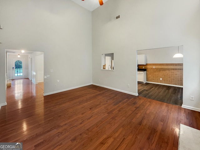 unfurnished living room with ceiling fan, a high ceiling, visible vents, baseboards, and dark wood finished floors