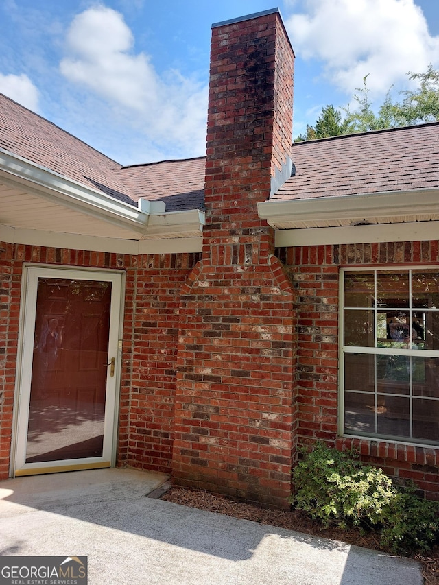 view of exterior entry with roof with shingles, a chimney, and brick siding