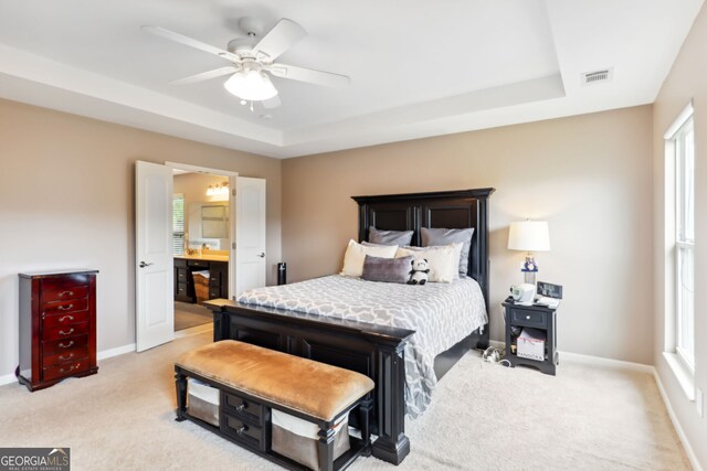 bedroom with a tray ceiling, light colored carpet, ensuite bathroom, and ceiling fan